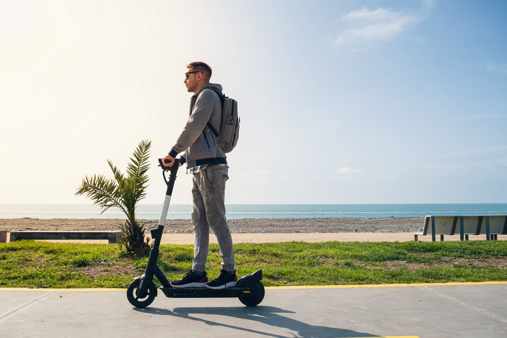 Young man traveler riding e-scooter along the sea coast and beach at sunny summer day. Male driving electric scooter